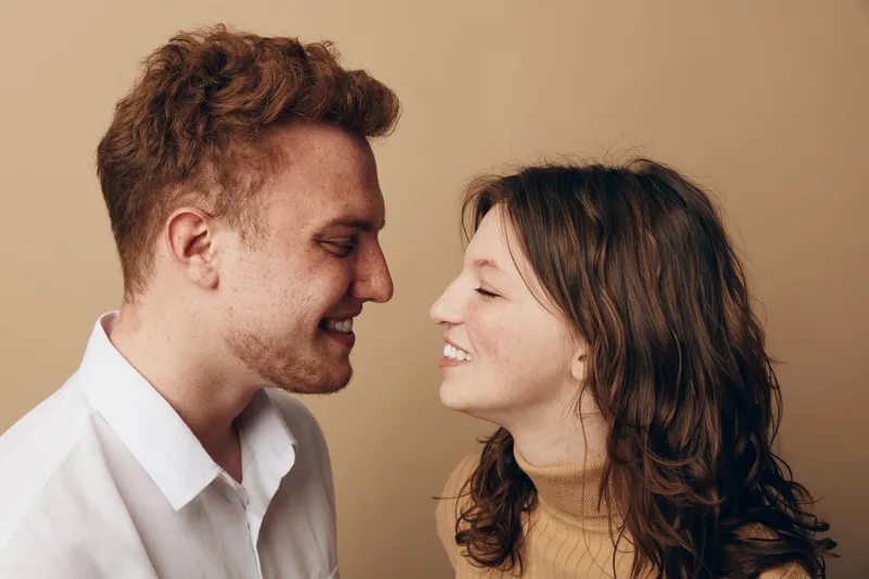 Smiling young couple with curly hair face each other closely, expressing affection and happiness. Warm beige background highlights their joyful and intimate moment together.