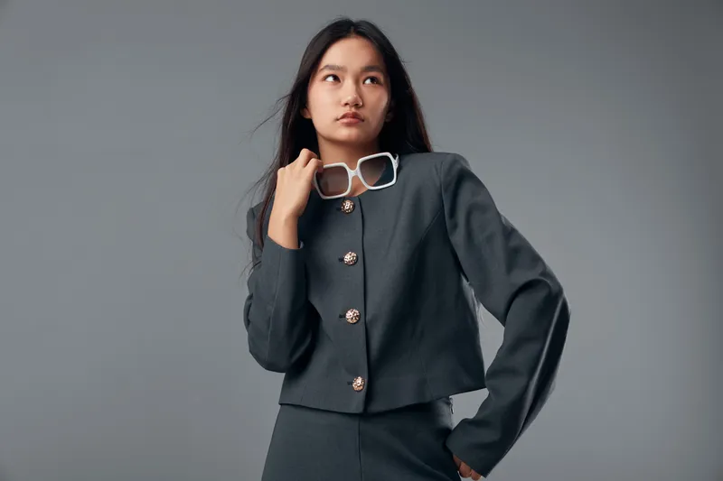 Young Asian woman posing in stylish dark gray outfit holding white sunglasses looking away in studio with plain gray background fashion model with long hair and serious expression.