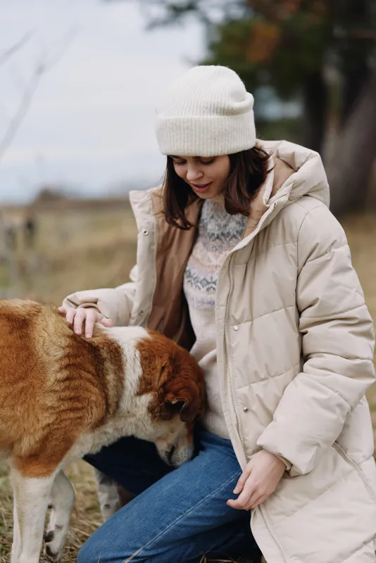 Woman in winter coat and beanie gently pets a dog in a field, outdoors, calm moment, soft light, nature surroundings, cozy scene of companionship
