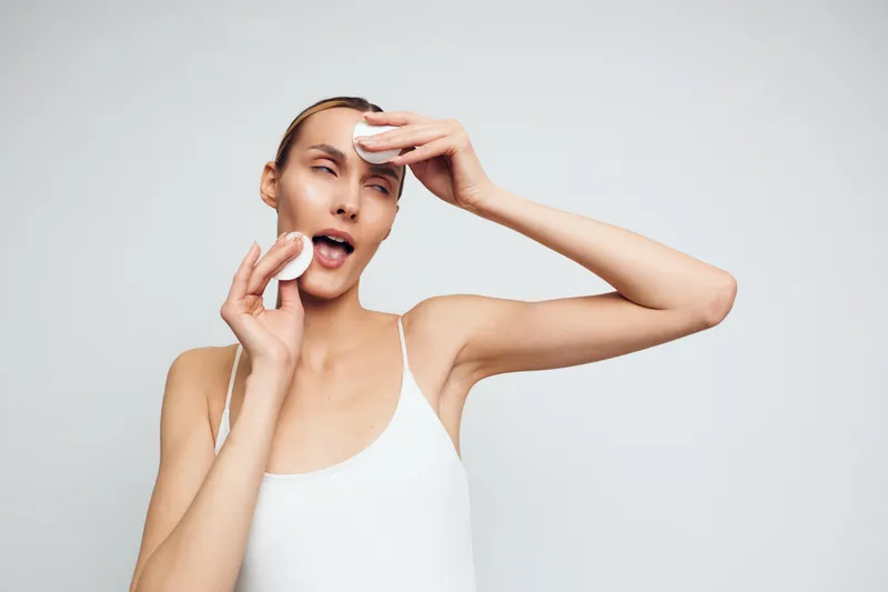 A young woman in a white tank top wipes her face with cotton pads, showcasing a skincare routine and a moment of self care in a bright studio.