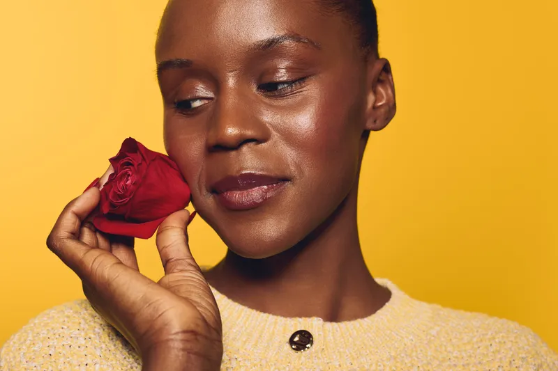 Close-up of a dark skinned African woman with smooth skin holding a red rose near her face on yellow background. Concept of beauty, skincare, self-care, and natural elegance.