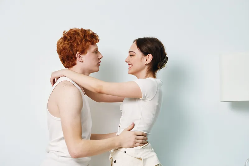Couple embracing indoors with casual clothes, young man and woman looking at each other, smiling and hugging in cozy bright room with white background and lamp.
