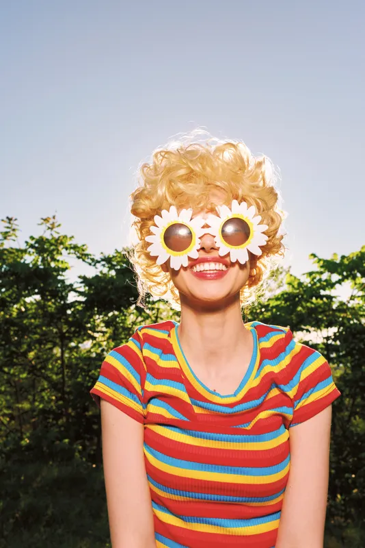 Cheerful woman wearing a rainbow striped shirt and oversized sunflower sunglasses stands outdoors on a sunny day, capturing a playful retro vibe with bright colors and warm glow.