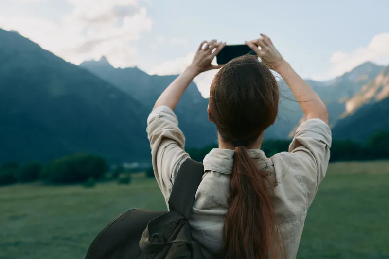 A woman outdoors with a backpack photographs distant mountains, capturing a moment of exploration and freedom. A calm lifestyle scene emphasizing nature, travel, and personal discovery.
