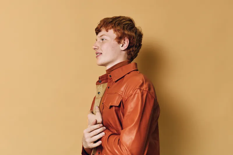 Redhead man wearing brown leather jacket looking sideways with neutral expression on beige background. Casual fashion, profile portrait, studio shot, young adult male, isolated, confidence.