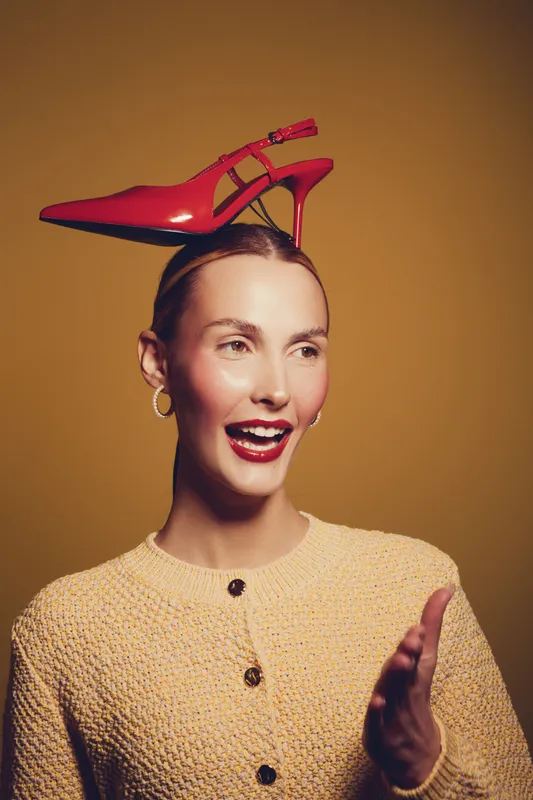 Smiling woman with red high heel shoe balanced on her head wears yellow cardigan and gold earrings. Studio portrait on brown background with playful and fashionable concept.