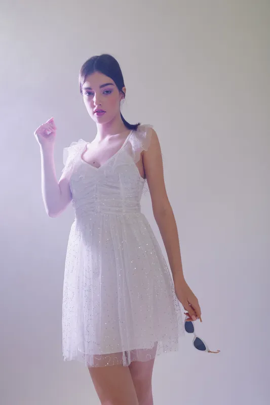 Young woman in a white dress poses holding sunglasses in a bright studio. The image focuses on beauty, fashion, and elegance with soft natural lighting.