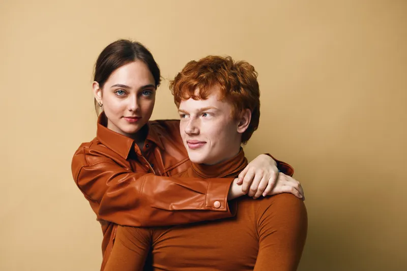 Couple with woman hugging man from behind on beige background. Young adults with casual clothes and affectionate pose for love and relationship concept.