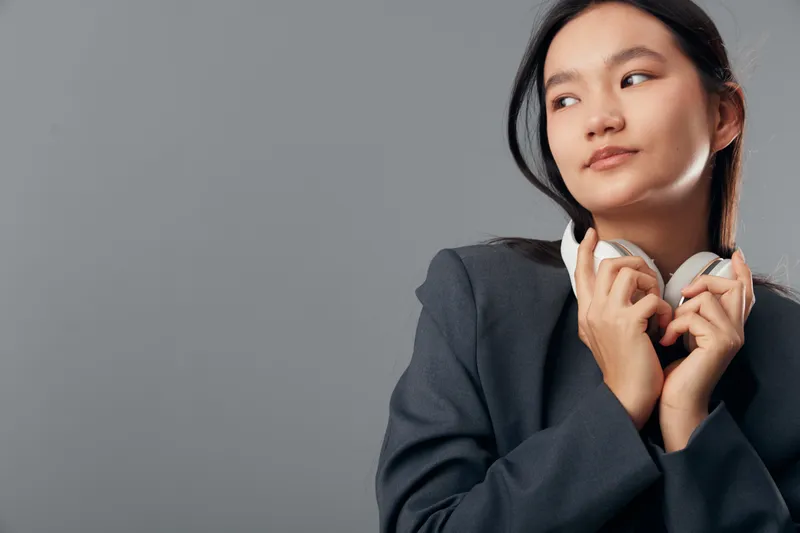 Young woman in business suit holding headphones looking away on grey background. Female professional with casual elegant style posing with audio device in studio portrait.