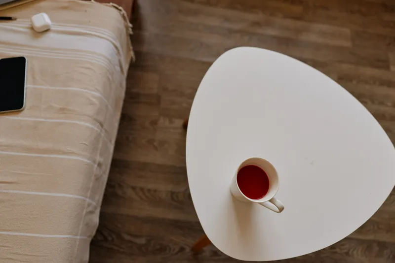 Cozy home scene with a cup of coffee on a small white triangular table beside a bed, warm morning light, relaxed mood, minimal decor, everyday lifestyle