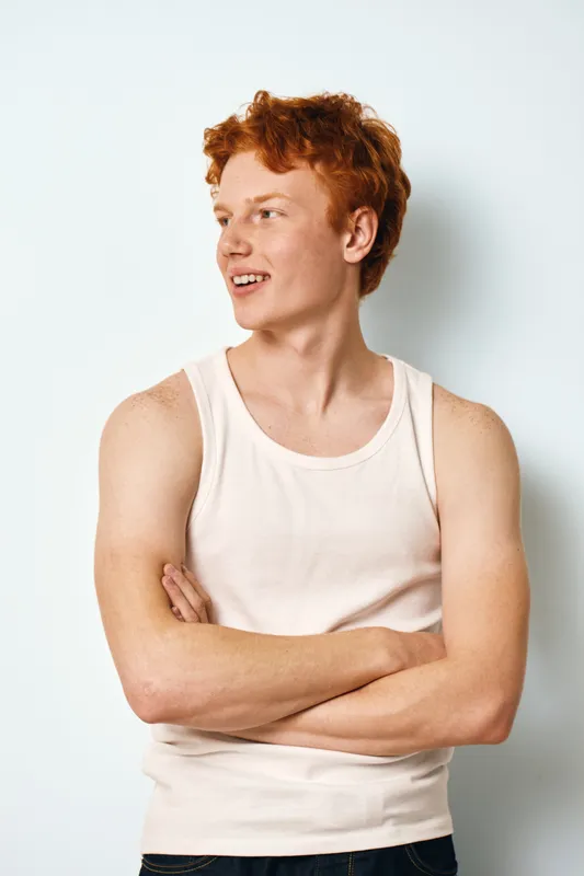 Young man with red hair and light skin wearing white tank top standing with crossed arms, smiling and looking sideways against plain background in casual style portrait.