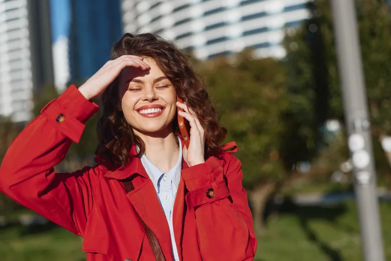 Young woman with curly hair smiles brightly while talking on a smartphone outdoors in a city park on a sunny day.