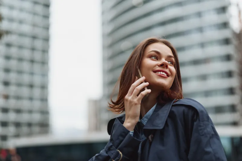 Smiling woman with brown hair wearing a blue jacket talks on a mobile phone in an urban outdoor setting, conveying communication, confidence, and contemporary lifestyle.