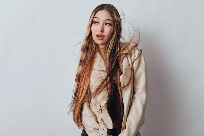 Woman with long hair and beige coat standing against plain wall for fashion and style in studio. Young female model with confident expression and natural look with casual outfit.