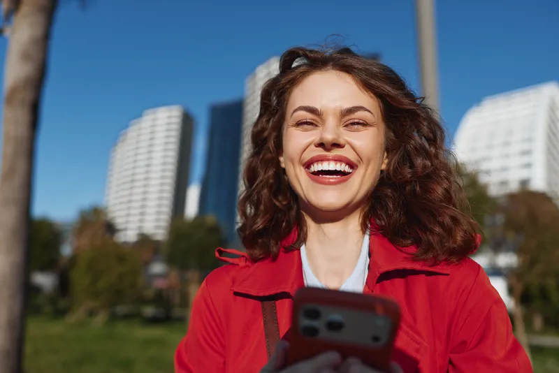 Smiling woman outdoors in a sunny city park, wearing a red jacket, holding a smartphone, capturing a vibrant moment of joy in an urban backdrop