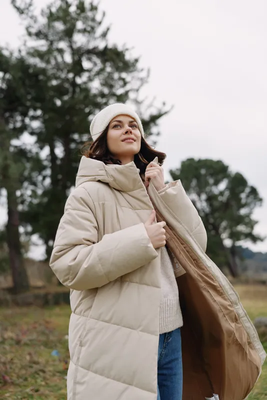 Woman wearing a beige puffer coat outdoors in autumn, gazing upward with a calm expression. Cozy knit sweater and casual fashion against a pine backdrop and open sky.