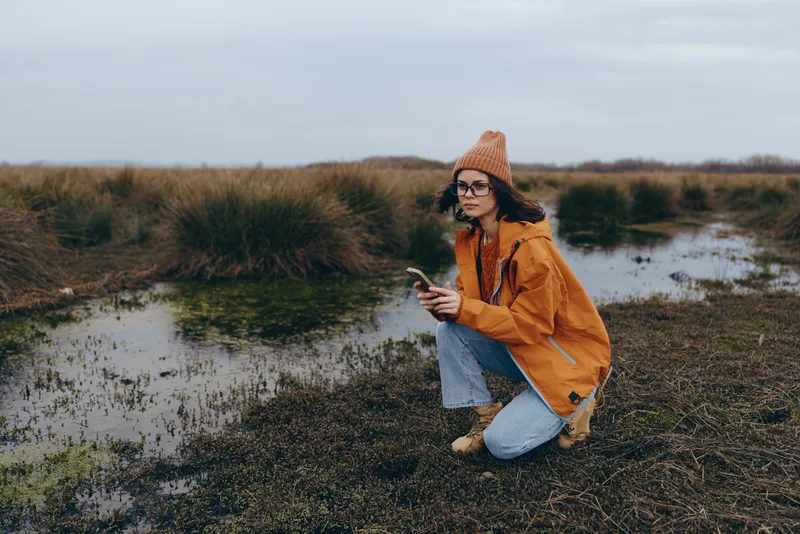 Woman in outdoor autumn nature lifestyle, kneeling by riverbank, wearing jacket, scarf and glasses, relaxed hand gesture, calm moment near water, natural light
