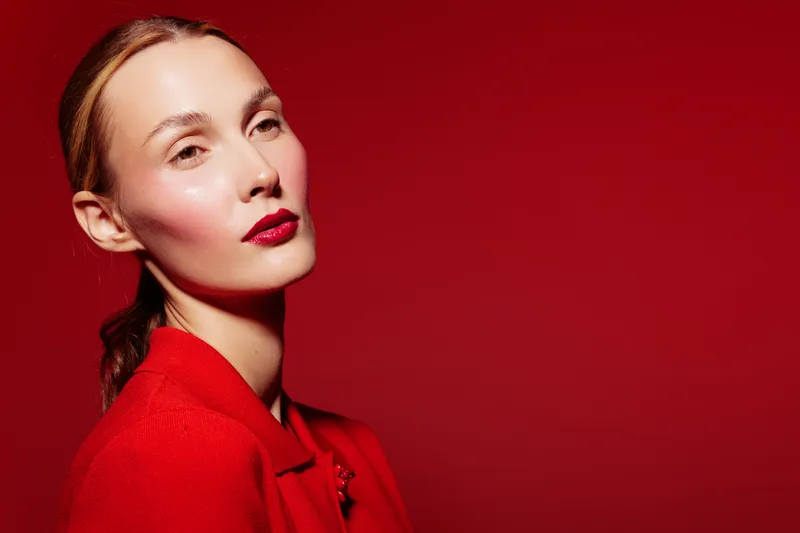 Portrait of confident woman with red lipstick and red jacket against red background. Studio lighting highlights her clear skin and elegant expression. Fashion and beauty concept.