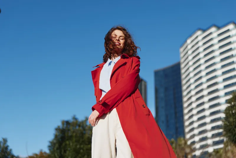 Confident model in a red coat standing outdoors with blue sky and tall buildings, showcasing bold street style and modern fashion for a dynamic editorial look.
