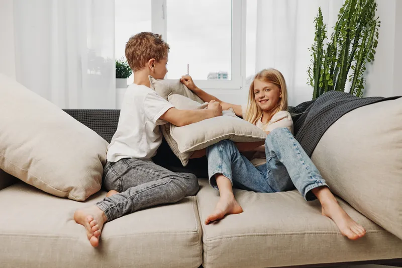 Two happy children having fun pillow fighting on beige sofa in bright living room. Casual clothes, relaxed atmosphere, and playful interaction between boy and girl at home.
