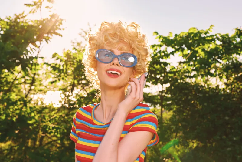 Cheerful woman outdoors, striped shirt, retro vibe, oversized sunglasses, laughing with head tilted, sunlit moment, relaxed pose, warm breeze, vivid colors, leafy background