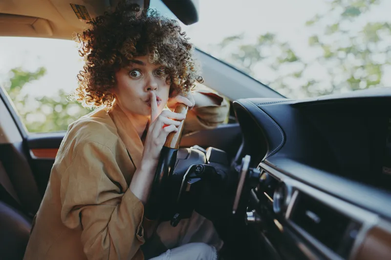 Cheerful woman inside a car, finger to lips in a playful hush, sunlit cabin with casual outfit, natural colors, candid lifestyle moment, relaxed atmosphere, warm light, soft breeze