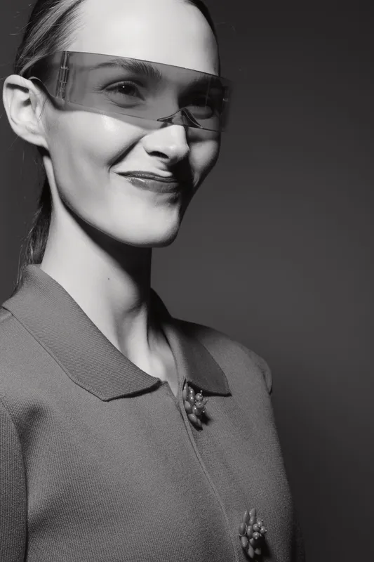 Smiling Caucasian woman with light skin wearing modern transparent glasses and a textured jacket, posing against a dark background in studio light.