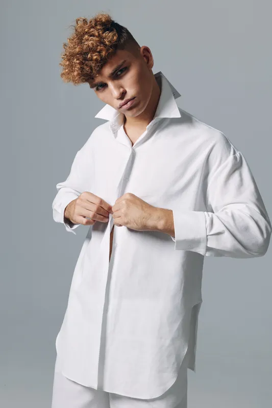 Young man with curly hair wearing a white oversized shirt standing in front of gray background, buttoning buttons, model posing indoors in modern fashion style studio.