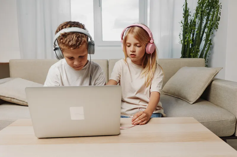 Two children wearing headphones use a laptop together at home on a beige sofa. The scene shows focus, learning, and digital entertainment in a cozy living room.
