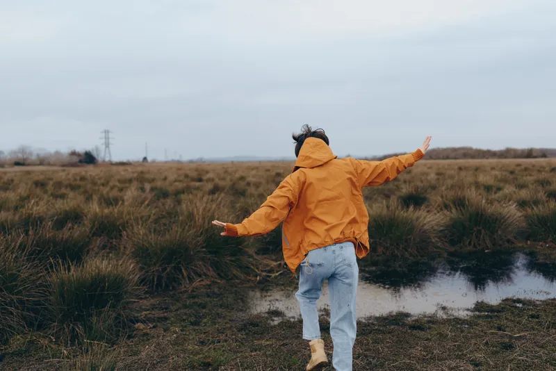 Woman lifestyle outdoors in autumn, wearing raincoat, walking through field, arms open, standing near puddle, cloudy sky, cozy adventure in nature, misty ground, boots, playful mood, full body