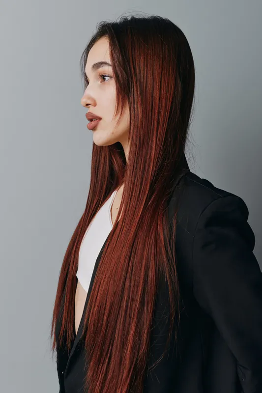 Young woman with long straight hair wearing black jacket and white top looking sideways on gray background. Profile portrait of female model with natural makeup and serious expression in studio.