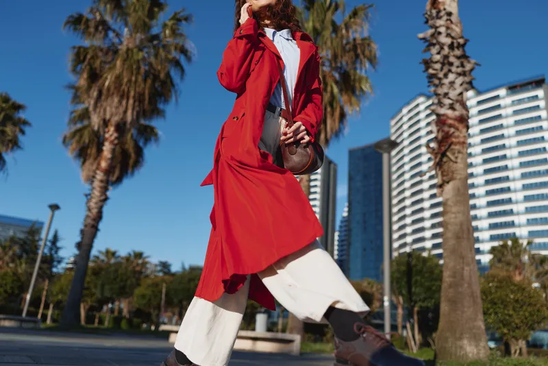 Young woman in a flowing red coat confidently walks through an urban park with palm trees and modern buildings under a clear blue sky.