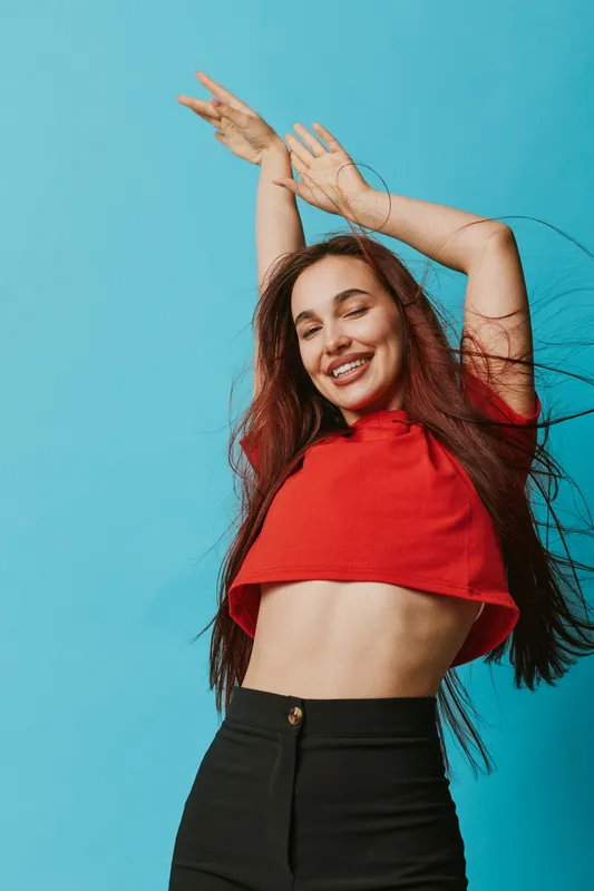 Young woman with long hair wearing red crop top and black pants dancing with arms raised against blue background. Happy female enjoying movement with closed eyes and smile.