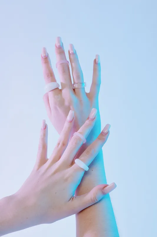 Close-up of female hands with long manicured nails and multiple rings on fingers. Soft blue and pink lighting highlights beauty and fashion concepts. Minimalist studio background.