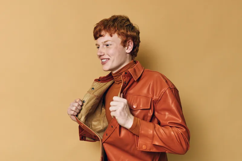 Young redhead man with curly hair wearing orange leather jacket and turtleneck smiling and posing confidently on beige background in studio lighting for fashion portrait.