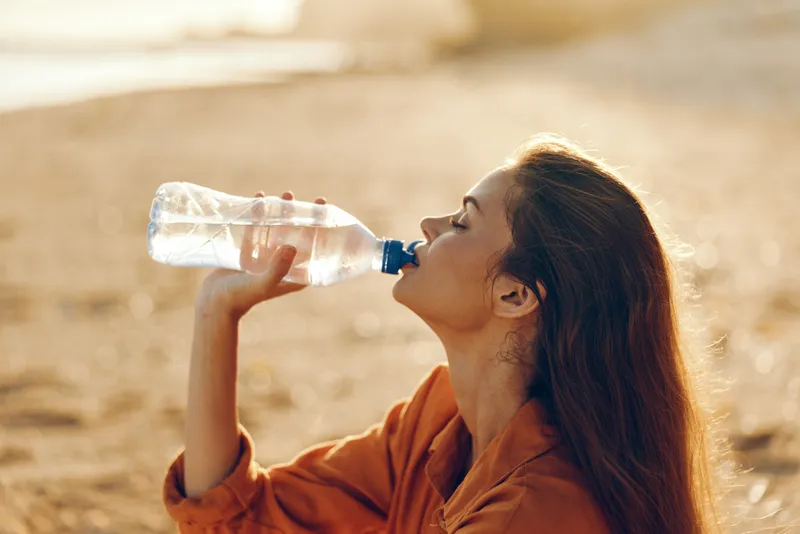 Young woman in a casual brown jacket drinks water from a bottle on a sunny beach, looking relaxed and focused as warm light glows over the sand.