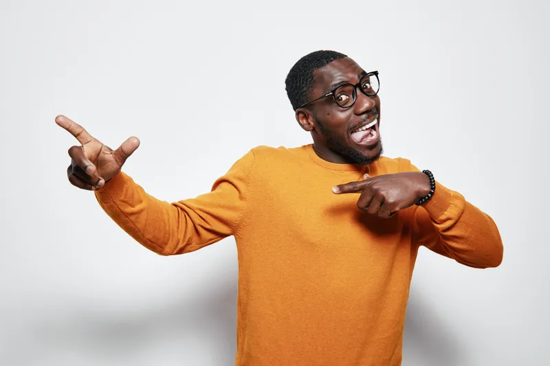 African American man with dark skin wearing glasses and orange sweater pointing fingers with excitement and joy on white background. Casual style and happy expression.