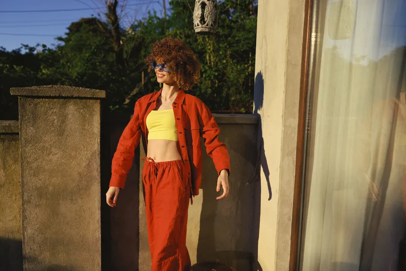 Young woman with curly hair in a yellow crop top and orange tracksuit stands on a sunlit balcony, relaxed, looking off to the side as a soft breeze brushes her outfit and the warm light highlights