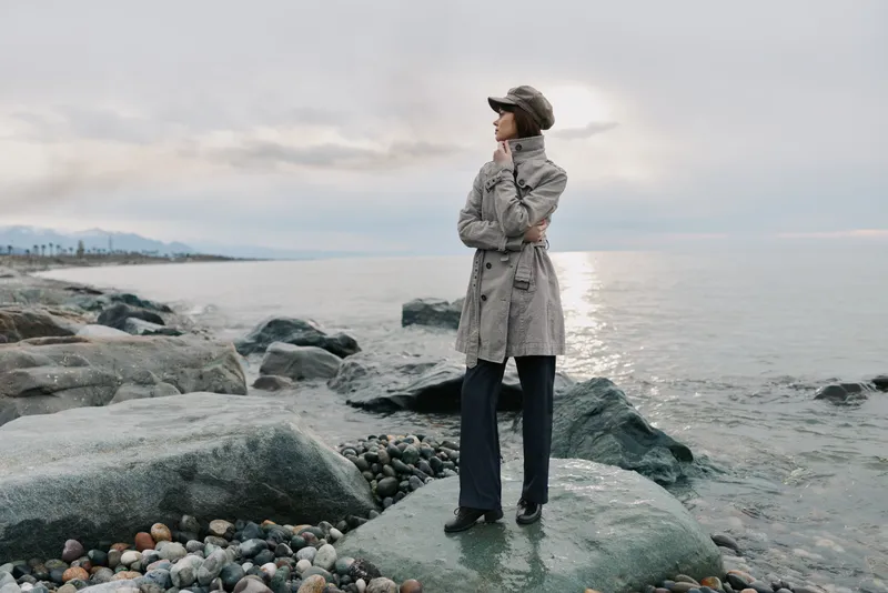 Woman in coat and cap stands on rocky shore looking at sea under cloudy sky. Concept for autumn fashion, outdoor lifestyle, and peaceful nature moments near water.