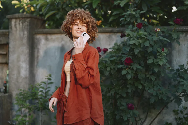 Casual lifestyle portrait of a smiling woman with curly hair talking on a smartphone in a sunny garden, wearing a rust coat and light top, natural relaxed mood