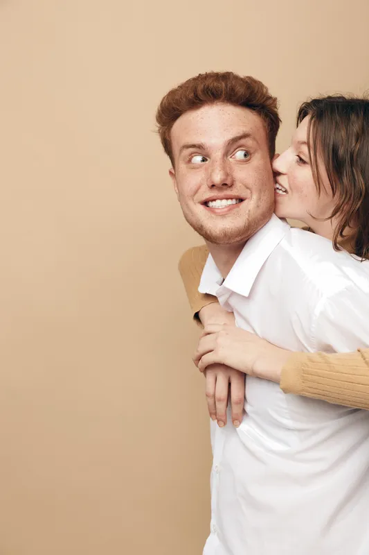 Happy young couple in casual clothes share joyful moment, woman hugging man from behind on beige background. Concept of love, friendship, relationship, and positive emotions.