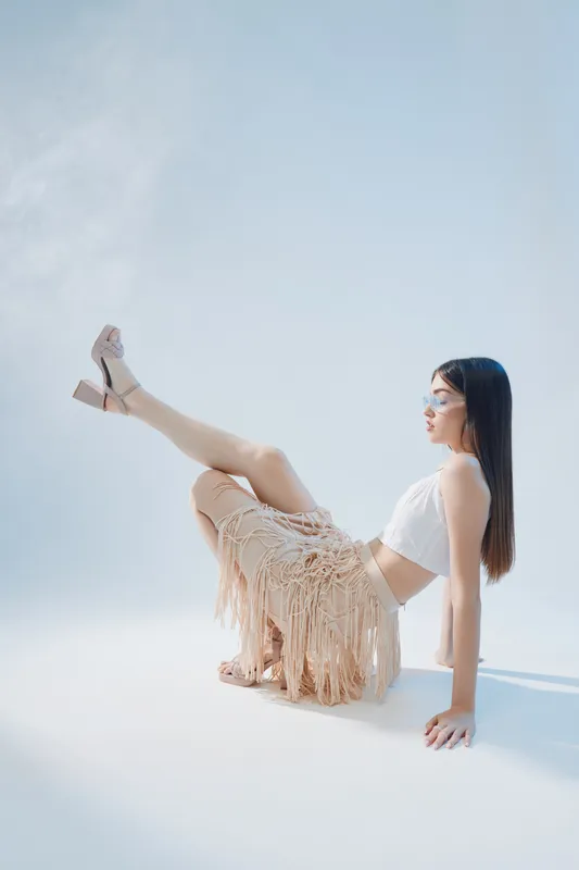 Young woman in a white top and fringed skirt poses elegantly on a light background. Concept of beauty, fashion, and modern style with soft natural lighting.