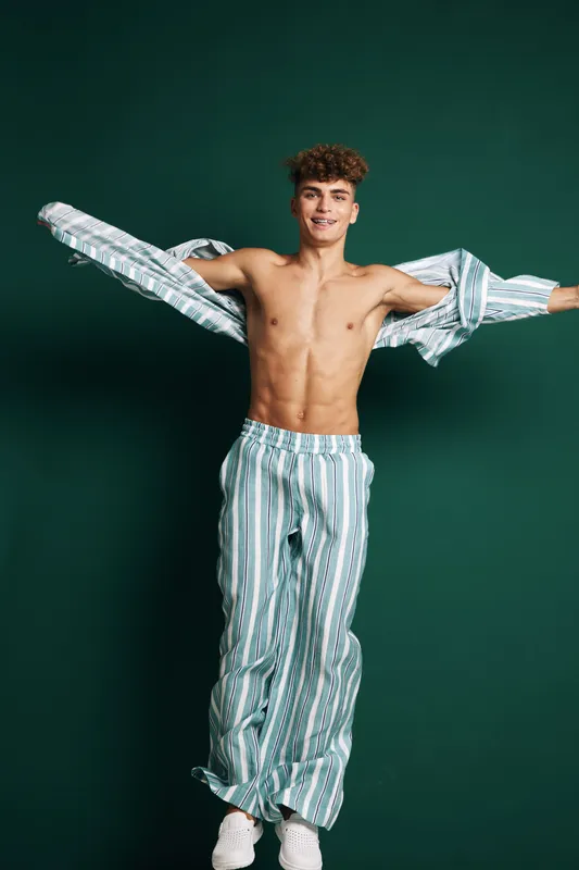 Young man with curly hair wearing striped pajamas and white sneakers smiling while opening his shirt against green background in studio. Casual, happy and relaxed pose.
