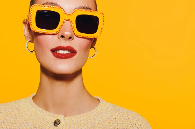 Stylish woman with orange sunglasses and red lipstick poses against bright yellow background. Fashion portrait with vibrant colors and confident expression. Concept of summer style.