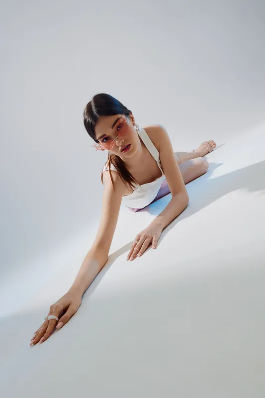 Young woman in white fashion outfit poses on white floor with pink sunglasses. Minimalistic beauty portrait with soft natural light and modern style concept.