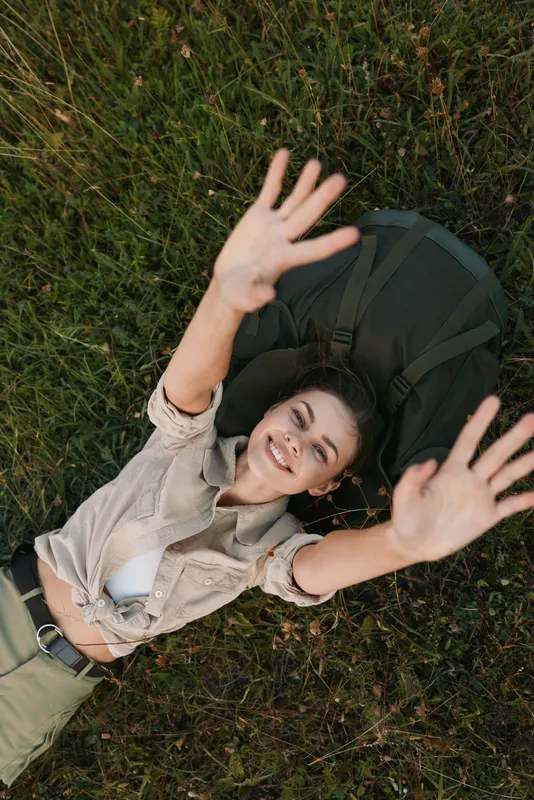 A smiling woman relaxes on grass outdoors, arms raised in a carefree pose. Casual outfit, natural light, and green surroundings convey a joyful, laidback lifestyle.