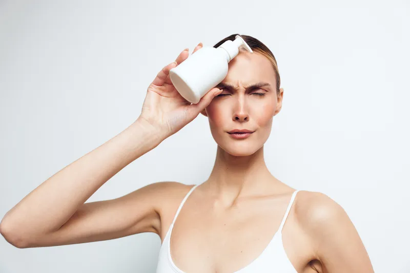 Young woman with clear skin holding a white cosmetic bottle near her forehead. Studio portrait on a white background. Concept of skincare, beauty, and self-care routine.