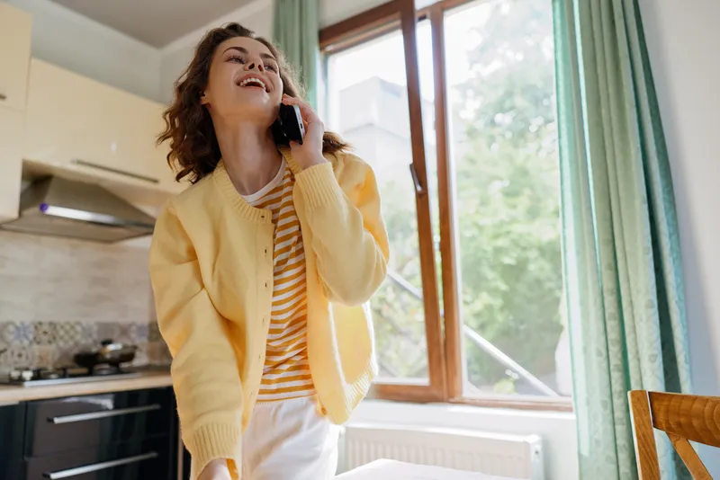 Cheerful woman indoors, wearing a yellow cardigan and striped shirt, talks on a phone in a bright kitchen, warm sunlight through a window, relaxed posture, candid lifestyle moment, cozy home
