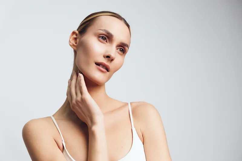 Woman portrait with a gentle neck touch, soft light highlights skin texture, calm serene mood, minimal studio setting, natural colors, graceful pose in a quiet moment