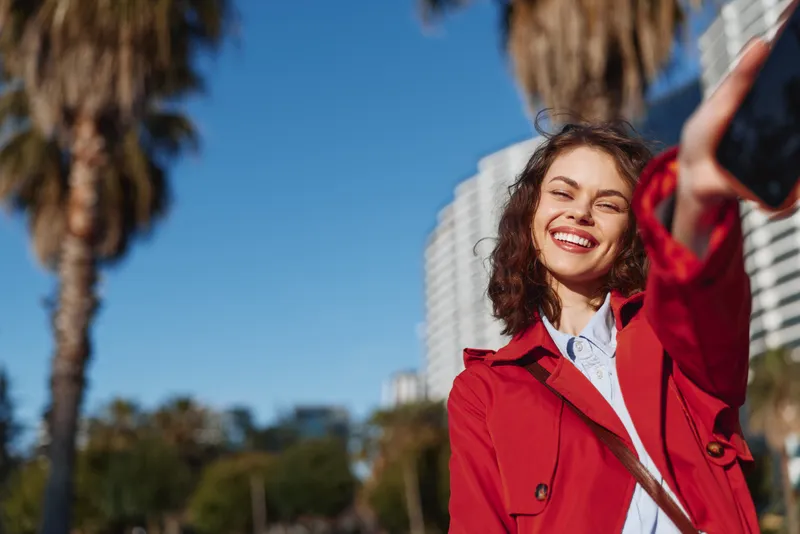 Young woman with curly hair in a red coat takes a selfie outdoors, smiling brightly as palm trees and high-rise buildings frame a sunny urban backdrop.